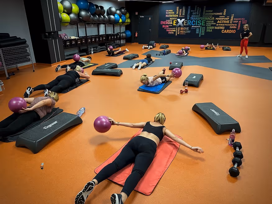 Grupowy trening fitness z instruktorem na sali – kobiety wykonują ćwiczenia w rytm muzyki.